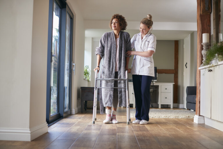 Mature Woman In Dressing Gown Using Walking Frame Being Helped By Female Nurse